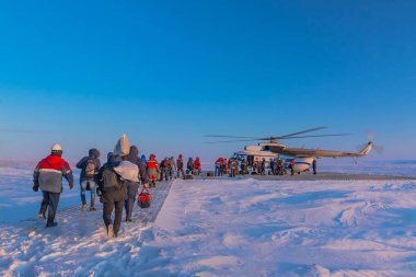 A polar aviation helicopter on a wooden heliport in the northern tundra, not far from a drilling oil well. The drillers and oil workers are dedicated and equipment is being loaded. Winter polar day. clipart