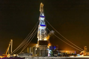 Gece aydınlatmalı petrol ve gaz sondaj kulesi. Kış kutup gecesi. Karla kaplı ekipmanların bulanık hatları.