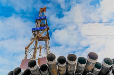 Petrol ve gaz kuyularını delmek için ön plandaki borularda. Arka planda bir sondaj kulesi var. Bulutlu mavi gökyüzü.