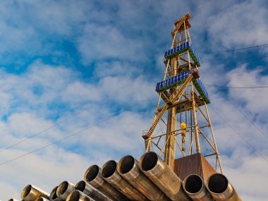 Petrol ve gaz kuyularını delmek için ön plandaki borularda. Arka planda bir sondaj kulesi var. Bulutlu mavi gökyüzü.