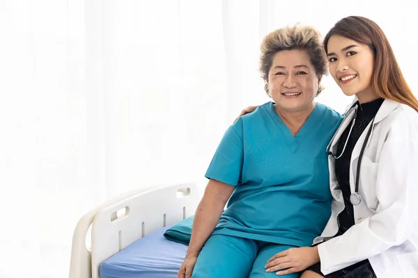 病院病棟での訪問中に制服を着た若い女性医師が抱き合って笑顔で高齢女性患者に — ストック写真