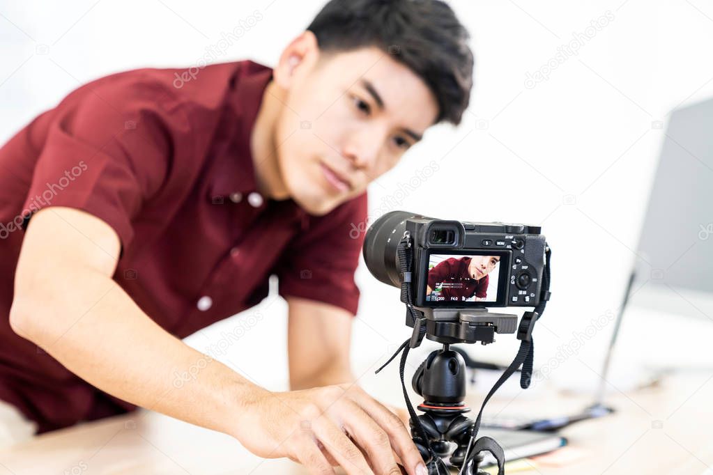 Young asian male blogger setting up camera for recording live vlog video tutorial session at home. IT blogging or vlogging, social media hobby broadcasting, or online learning course concept. Focus on camera
