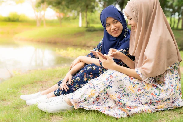 若い大人のティーンイスラム教徒のイスラム教徒のタイの女性大学の学校で湖や緑の公園の横にソーシャルメディアでスマートフォンやデジタルタブレットを使用して — ストック写真