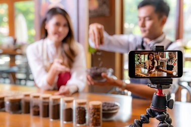Genç yetişkin Asyalı girişimci kafe kahve dükkanının video bloğu ve kafedeki akıllı telefonuyla çevrimiçi pazarlama için sosyal medyada yaşıyor. Küçük işletme oluşturma ve tüketim kavramının başlangıcı için kullanılacak. 