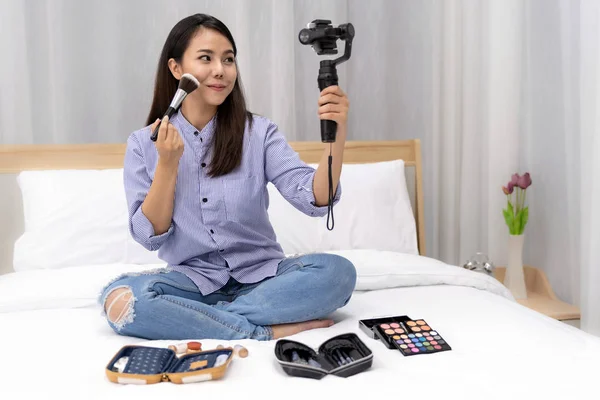 Young beautiful Asian woman beauty vlogger or blogger recording live how to make up tutorial to share on social media using Gimbal Stabilizer on mobile phone. Using for vlog social media influencer concept.