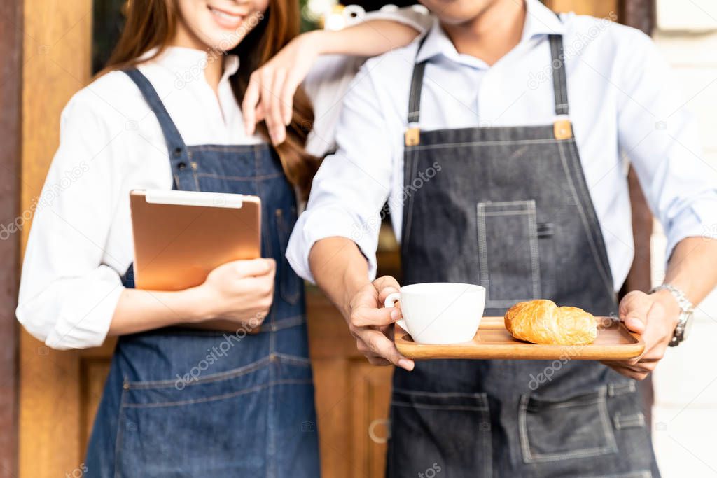 Closeup Asian young adult owners of Small business cafe holding coffee set and digital tablet in front of coffee retail shop. Using as startup SME and consumerism concept.