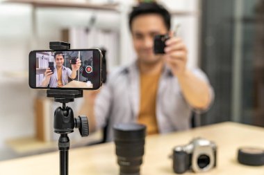 Genç Asyalı erkek BT blogcusu ve blog yazarı dijital kamera teknolojisi hakkında canlı yayında cep telefonunu kullanarak video kaydı yapıyor. Çevrimiçi sosyal medya kavramı. Kameraya odaklan..