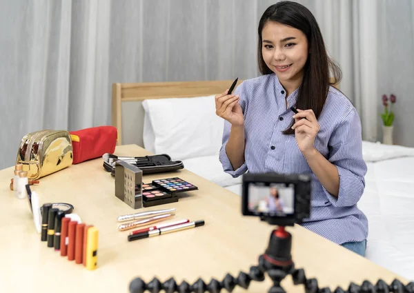 Young beautiful Asian woman beauty vlogger blogger recording live make up tutorial share on social media using digital camera on tripod. Using for vlog social media influencer concept. Focus on camera.