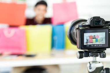 Genç Asyalı erkek çevrimiçi ticari blog yazarı renkli alışveriş çantasıyla canlı video kaydediyor ve evde satış ve teftiş için hazırlanıyor. Çevrimiçi olarak sosyal medya kavramını etkiliyor. Kameraya odaklan.