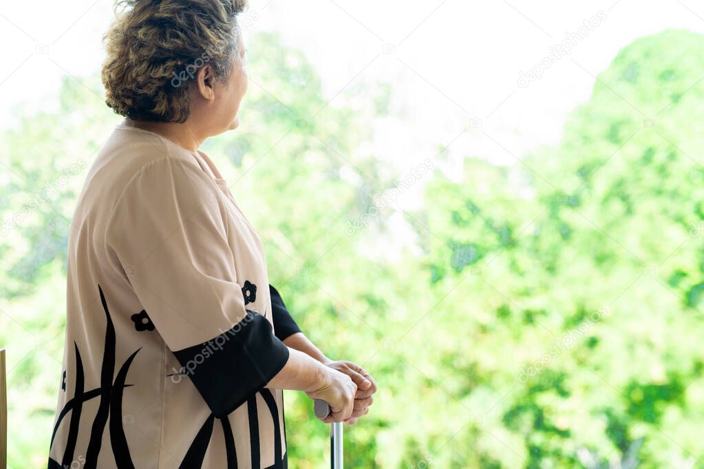 Elderly senior female using walking stick or walking cane staff standing beside window in domestic living room - recovery and rehabilitation concept