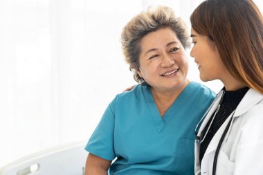 Üniformalı genç bayan doktor hastane koğuşunda yaşlı kadın hastalara sarılıp gülümsüyor.