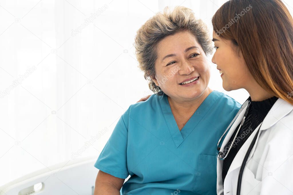 Young female doctor in uniform hugging and smiling to old elderly woman patient during a visit in hospital ward