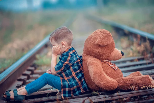 Un petit garçon avec un gros ours en peluche est assis sur la voie ferrée. La solitude et la tristesse d'un enfant. Le problème des enfants abandonnés — Photo