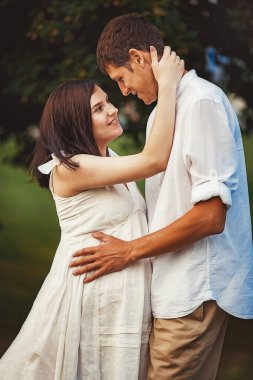 Bir erkek doğada hamile karısına nazikçe sarılır. Beyaz elbiseli mutlu evli çift birbirlerine bakıp gülümsüyor. Bir çocuğun doğmasını bekliyorum..