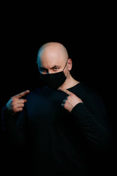 A bald man in a black virus mask on a black background. Protection and prevention from infection. Portrait of a serious man in a dark key