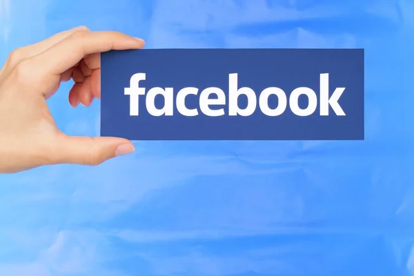 Ręką trzyma Facebook logo na tle niebieski papier — Zdjęcie stockowe
