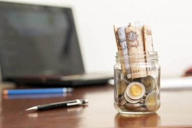 Coins and Banknotes in Jar, Egyptian Pounds, on Wooden Desk with Laptop and Pens clipart