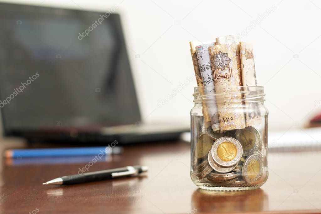 Coins and Banknotes in Jar, Egyptian Pounds, on Wooden Desk with Laptop and Pens