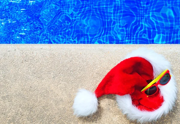 Κόκκινο Χριστουγεννιάτικο καπέλο και γυαλιά ηλίου κοντά στην πισίνα. Θέση για το κείμενο. — Φωτογραφία Αρχείου