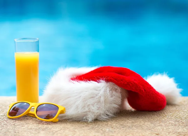 Χριστουγεννιάτικο καπέλο, γυαλιά ηλίου και χυμό πορτοκάλι κοντά στην πισίνα. — Φωτογραφία Αρχείου