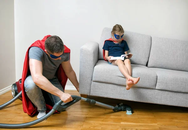 Ordinary day of superhero family. Dad vacuuming, daughter reading a book.