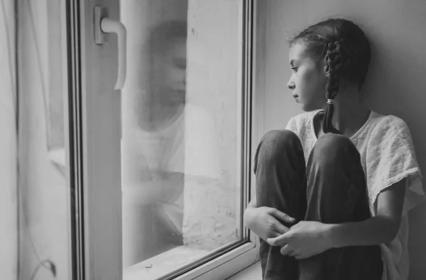 Sorgliga lilla flicka sitter nära fönstret. Svart och vitt. — Stockfoto