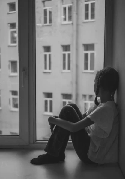 Sorgliga lilla flicka sitter nära fönstret. Svart och vitt. — Stockfoto