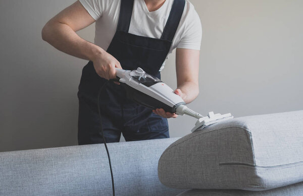 Man in uniform cleaning fabric of the sofa with dry steam cleaner.