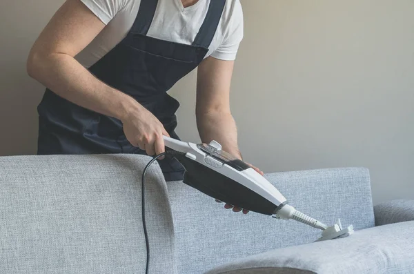 Man in uniform cleaning fabric of the sofa with dry steam cleaner. — Stock Photo, Image