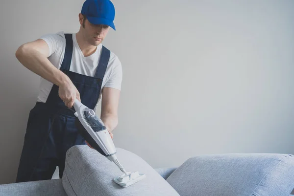 Man in uniform cleaning sofa with dry steam cleaner. Place for text. — Stock Photo, Image