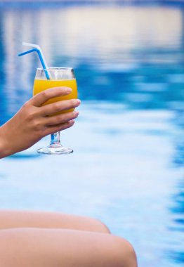 Havuzun kenarında oturan ve elinde portakal suyu olan bir kadın..