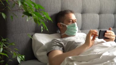 Yatakta cep telefonu kullanan maskeli hasta bir adam..
