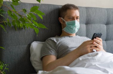 Yatakta cep telefonu kullanan maskeli hasta bir adam. Ev karantina konsepti.