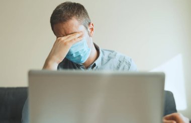Tıbbi maskeli yorgun adam içeride bilgisayar üzerinde çalışıyor..