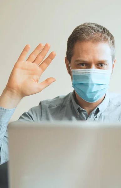 Man in medical mask greets via video calling indoors.