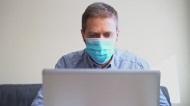 Tıbbi maskeli yorgun adam içeride bilgisayar üzerinde çalışıyor..