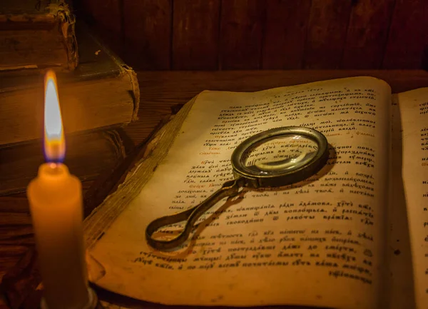 An open ancient prayer book and a magnifying glass on its open pages under the light of a candle