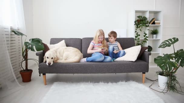 Dispositivos na vida das crianças. irmãozinho e irmã jogando no smartphone no jogo, cão triste deitado lado a lado no sofá . — Vídeo de Stock