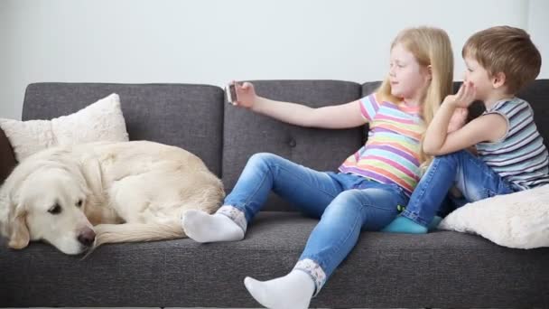 Dispositivos en la vida de los niños. hermano pequeño y hermana hacer selfie en un teléfono inteligente, perro triste se encuentra en el sofá . — Vídeo de stock