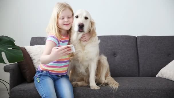 Dispositivos en la vida de los niños. Una niña pequeña hace selfie con su perro en un smartphone. Ubicación sofá en la sala de estar . — Vídeo de stock