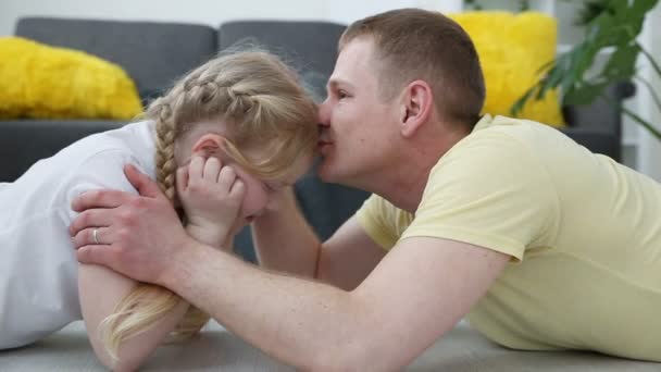 Vård för barn i familjen. Pappa pussar och kramar en liten dotter, liggande på golvet i vardagsrummet. — Stockvideo