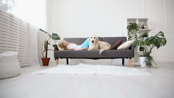 Cuidado de mascotas. Una chica rubia acaricia a su perro con amor en la sala de estar. feliz golden retriever en la familia . — Vídeo de stock