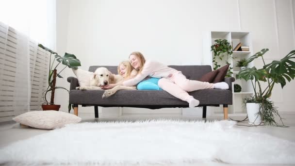 Livet av husdjur i familjen. mamma och dotter smekte och krama en golden retriever på soffan i vardagsrummet. — Stockvideo