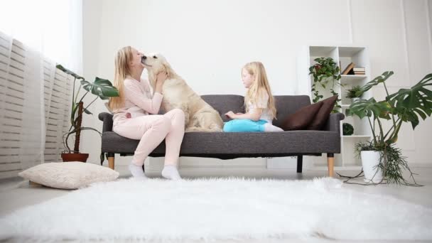 Życie zwierząt domowych w rodzinie. Mama i córka pogłaskał i przytulić golden retriever na kanapie w salonie. — Wideo stockowe