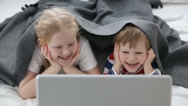 Computers in het leven van de moderne kinderen. kleine broer en zus kijken cartoons op laptop, liggend op bed in de slaapkamer. — Stockvideo