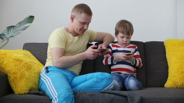 Dispositivos en la vida de una familia moderna. papá con un pequeño hijo jugando juegos en teléfonos inteligentes, sentado en el sofá en casa . — Vídeos de Stock