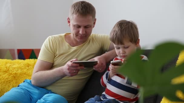 Enheter i livet för en modern familj. Pappa med en liten son som spelar spel på smartphones, sitter i soffan hemma. — Stockvideo