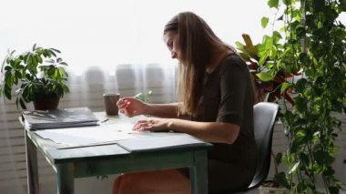 evde çalışıyorum. genç güzel freelancer kadın evde pencere kenarındaki masada çalışıyor. yaşayan bitkiler ile modern ekolojik iç.