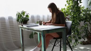 evde çalışıyorum. genç güzel freelancer kadın evde pencere kenarındaki masada çalışıyor. yaşayan bitkiler ile modern ekolojik iç.