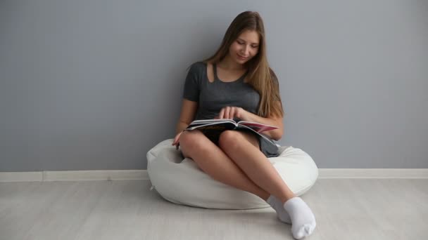 Una chica de aspecto europeo lee una revista en casa en una bocanada junto a la pared gris — Vídeo de stock